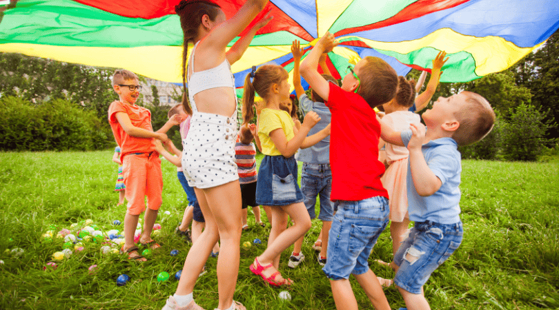 A group of children laughing and playing under a large, colorful parachute during a summer activity.