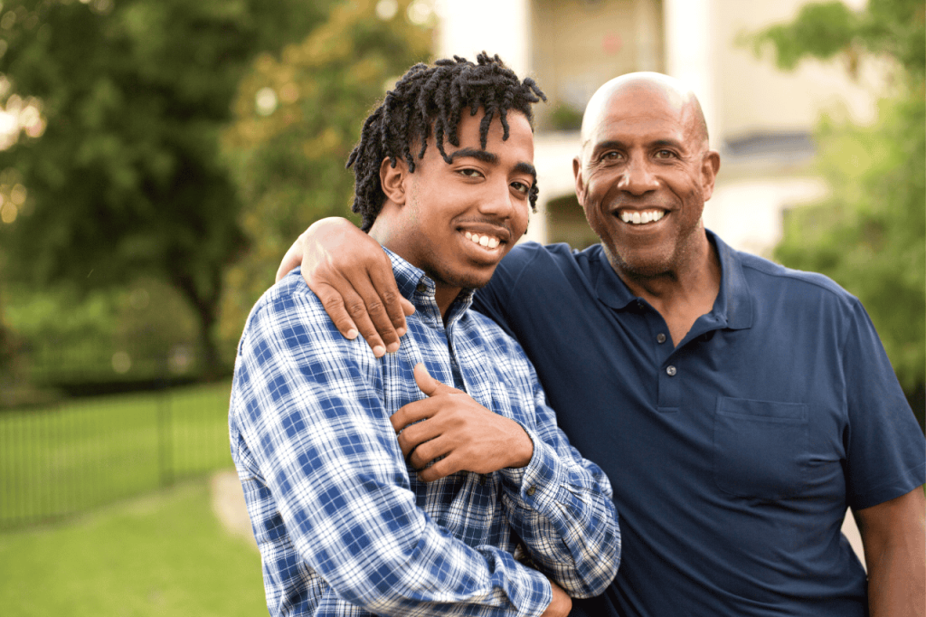 Strengthening Parent-Teen Bonds through Family Therapy – Lamplight Counseling A man with his arm around his older teenage son, both smiling and standing outside in a natural setting.